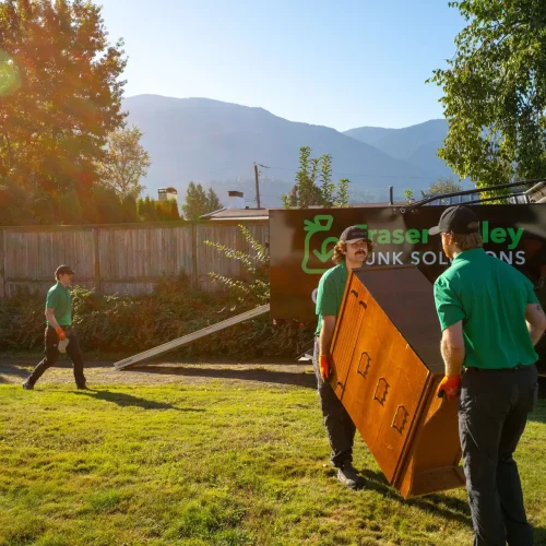 estate-downsizing-services two men wearing green bring a dresser to a 17.5 cubc yard truck for disposal on a nice day